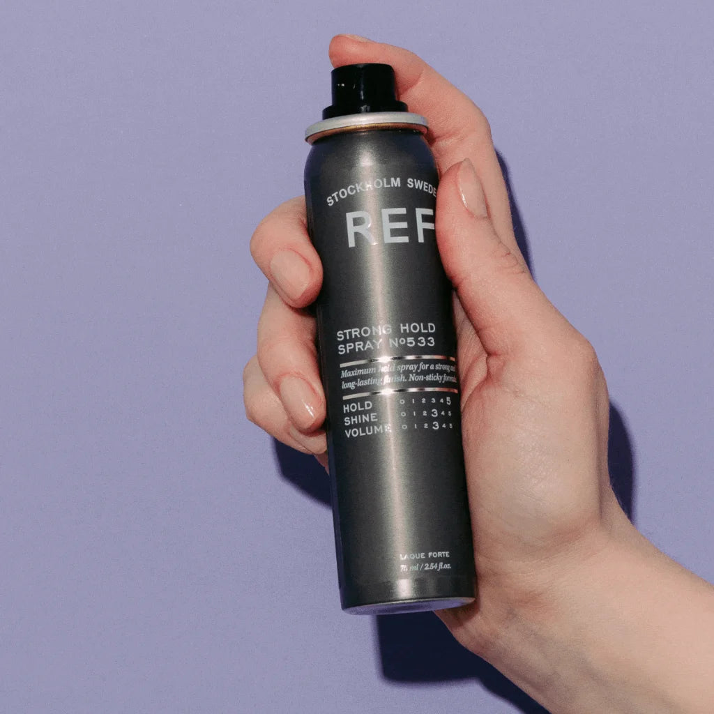 A hand holding a can of REF Stockholm Sweden Strong Hold Spray No. 33 hair styling product against a solid lavender background. MANEPRINT